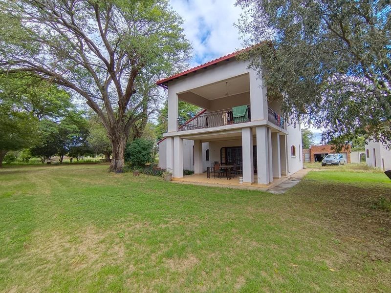 Rear view of a two-story farmhouse featuring a covered ground-floor patio and an upper-level balcony set within a spacious garden.