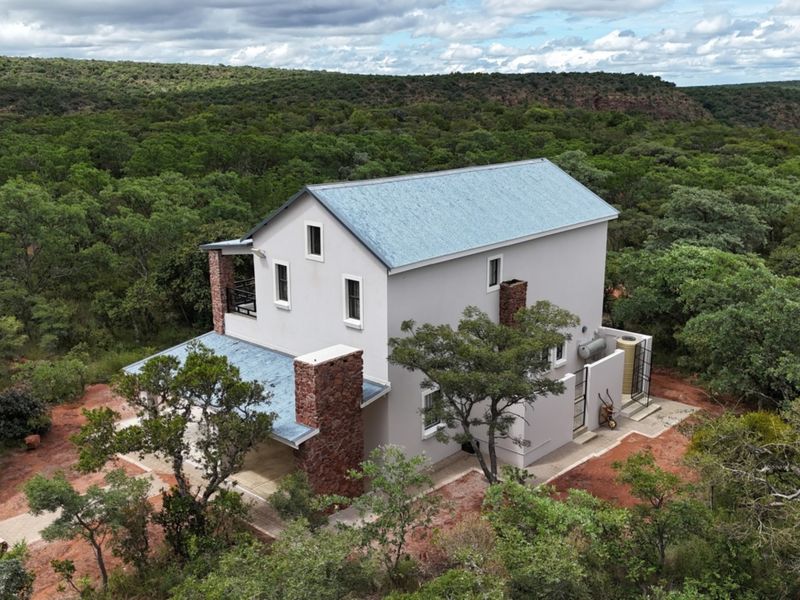 Aerial view of a two-story house featuring a metal roof, stone chimney accents, and a surrounding natural bushveld landscape.