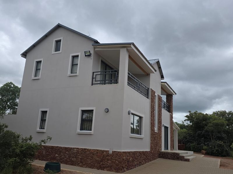 A modern two-story house exterior featuring stone cladding, a balcony with metal railings, and a paved walkway set against a natural landscape.