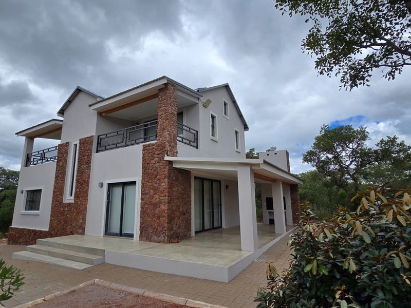 A modern two-story house featuring stone cladding, a covered patio with a built-in braai, and an upper-level balcony overlooking the surrounding natural landscape.
