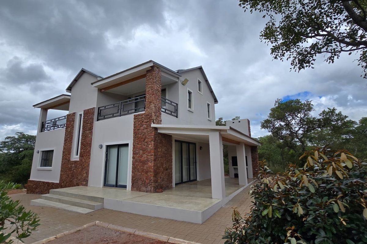 A modern two-story house featuring stone cladding, a covered patio with a built-in braai, and an upper-level balcony overlooking the surrounding natural landscape.