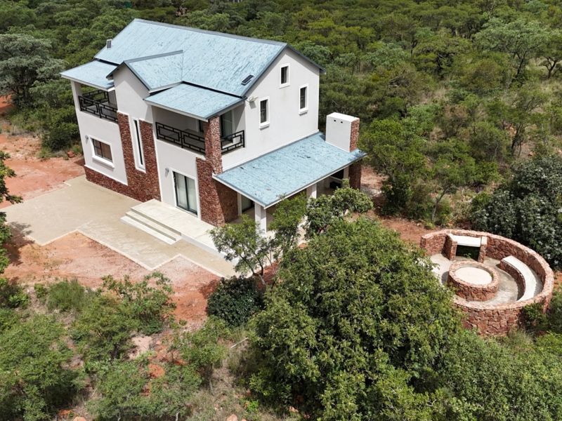 Aerial view of a two-story house featuring stone cladding, metal roofing, a balcony, and a circular stone-built fire pit area in a natural setting.