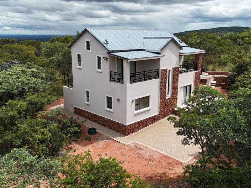 Aerial view of a two-story house featuring stone cladding, a metal roof, and a paved driveway, set within a natural bushveld landscape.