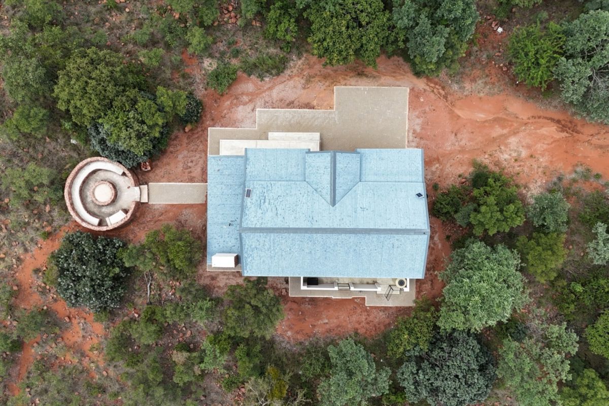 Aerial view of a residential property featuring a metal roof, paved patio area, and a circular outdoor fire pit set within a natural landscape.