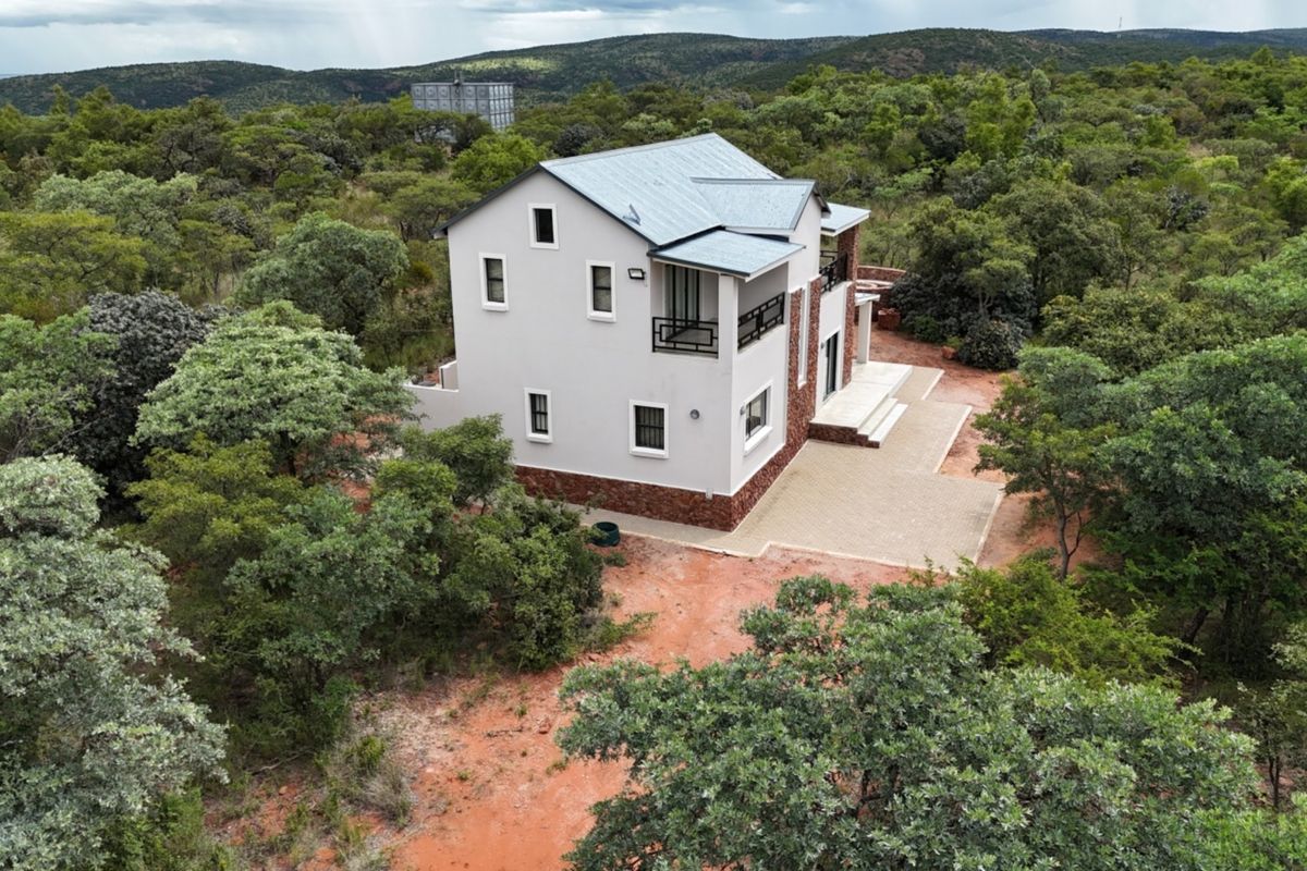Aerial view of a modern two-story house with stone cladding and metal roofing, situated in a natural bushveld setting with a paved patio area.