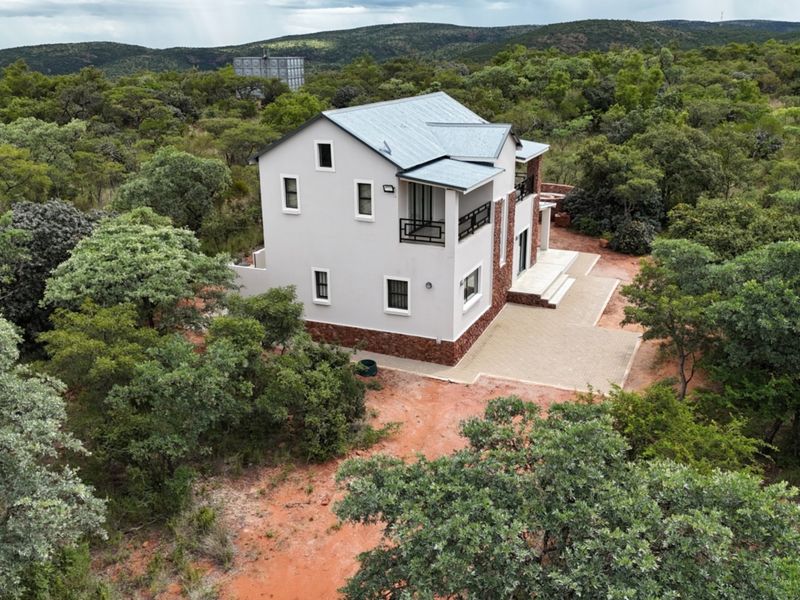 Aerial view of a modern two-story house with stone cladding and metal roofing, situated in a natural bushveld setting with a paved patio area.
