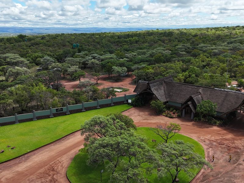 Aerial view of a residential property entrance featuring a thatched roof structure, perimeter wall, and expansive landscaped grounds set within a natural bushveld environment.