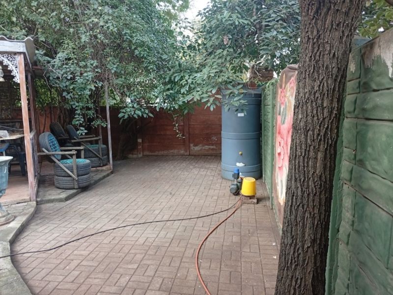 A paved garden area featuring a water storage tank, pump, and a covered patio structure, enclosed by a precast concrete boundary wall.