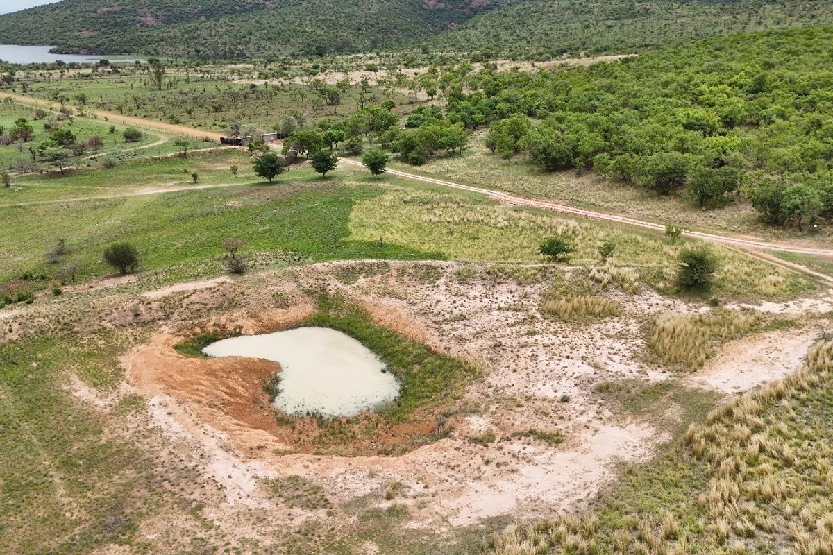 Aerial view of a vacant residential land plot featuring a natural pond, native vegetation, and an unpaved access road within a scenic landscape.