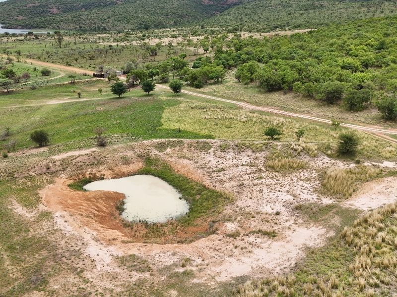 Aerial view of a vacant residential land plot featuring a natural pond, native vegetation, and an unpaved access road within a scenic landscape.