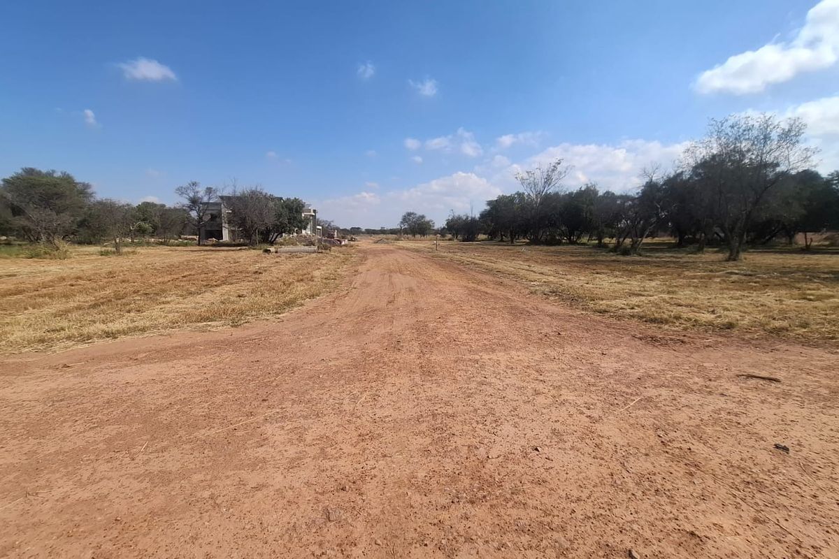 A view of an unpaved access road leading through a vacant residential plot with surrounding natural vegetation.