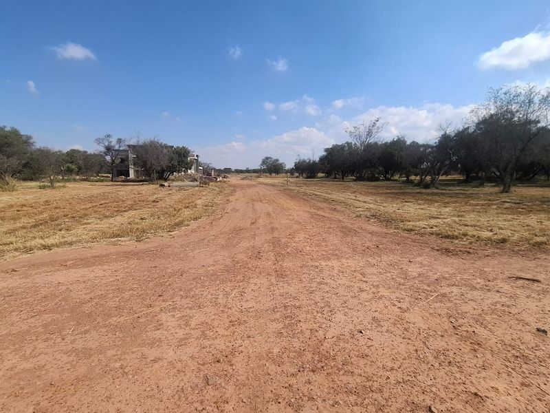 A view of an unpaved access road leading through a vacant residential plot with surrounding natural vegetation.