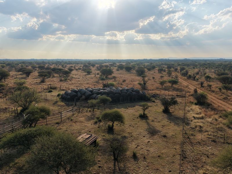 An aerial view of the expansive farm landscape featuring natural vegetation, a dirt access track, and fenced livestock enclosures under a dramatic sky.