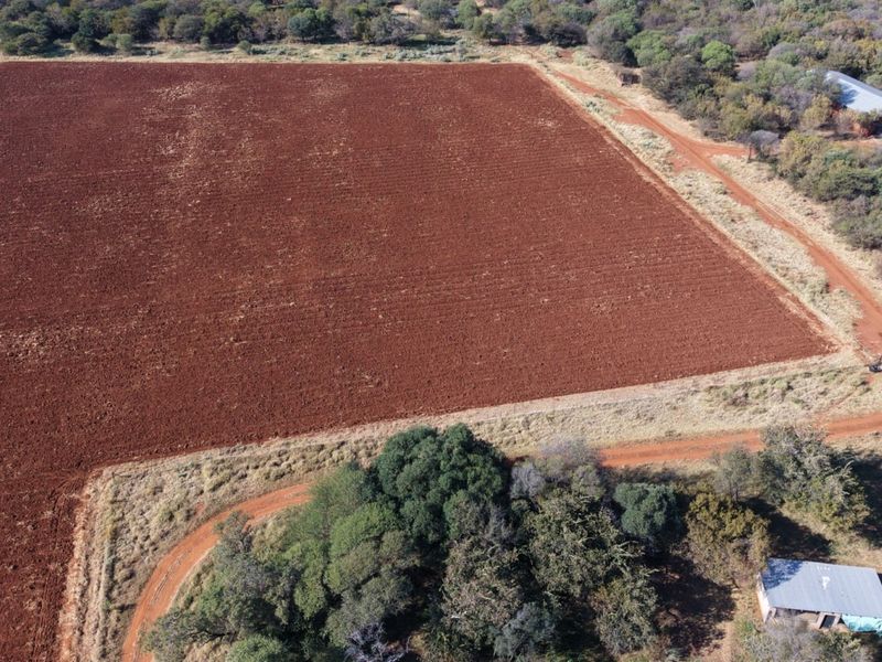 Aerial view of a large, tilled agricultural field on a farm, featuring surrounding natural vegetation, dirt access roads, and small outbuildings.