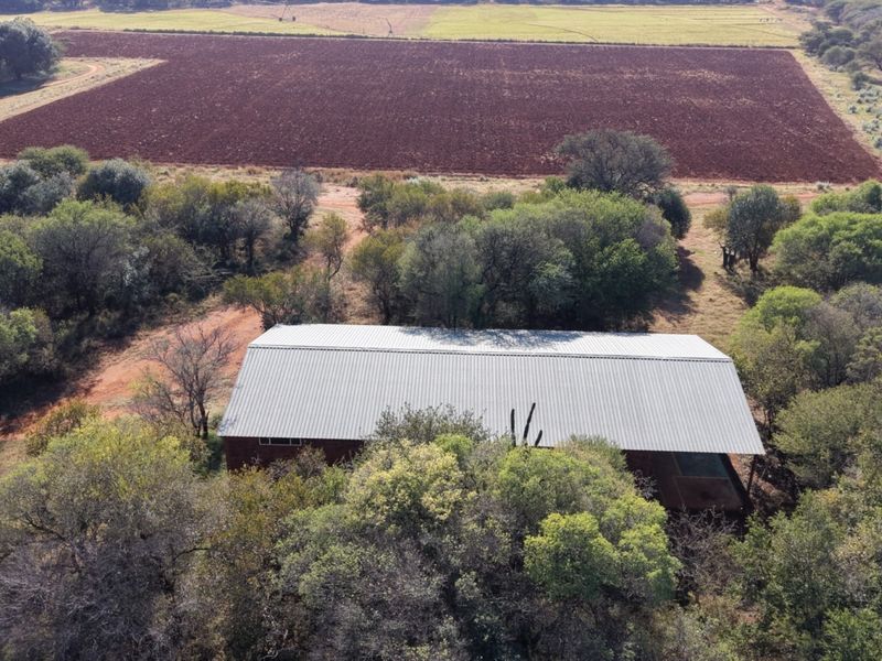 Aerial view of a farm property featuring a large structure with a corrugated metal roof surrounded by trees and adjacent ploughed agricultural fields.