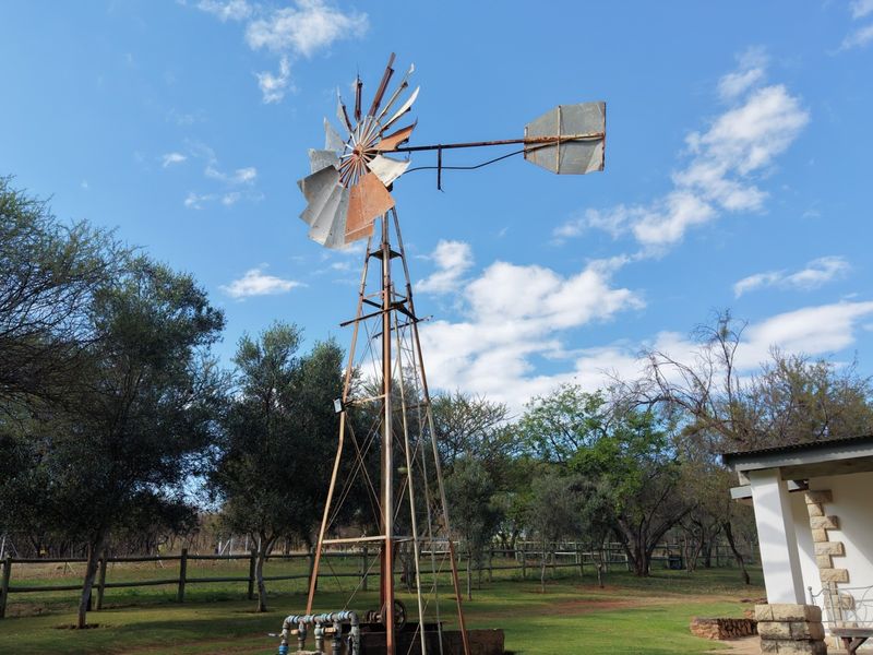 A traditional farm windmill and water pump infrastructure situated in a landscaped garden area with surrounding fencing.