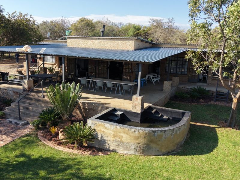 Aerial view of a farm-style residence featuring a large covered patio, built-in braai, and a raised stone water feature set within a landscaped garden.