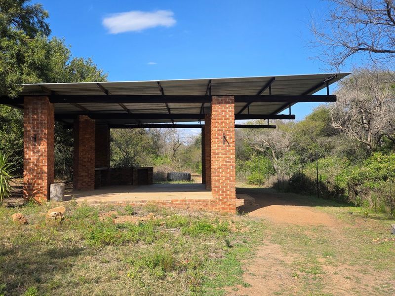 An open-air brick and metal roof entertainment area featuring a built-in braai, set within a natural bushveld garden environment.