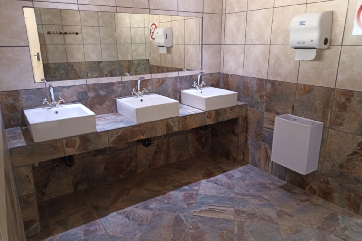 A functional communal bathroom featuring a triple vanity with vessel sinks, tiled surfaces, and wall-mounted fixtures.