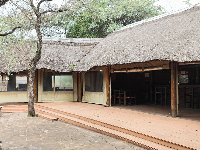 Spacious outdoor entertainment area featuring a large thatched roof, timber decking, and roll-up canvas blinds, set within a natural garden environment.