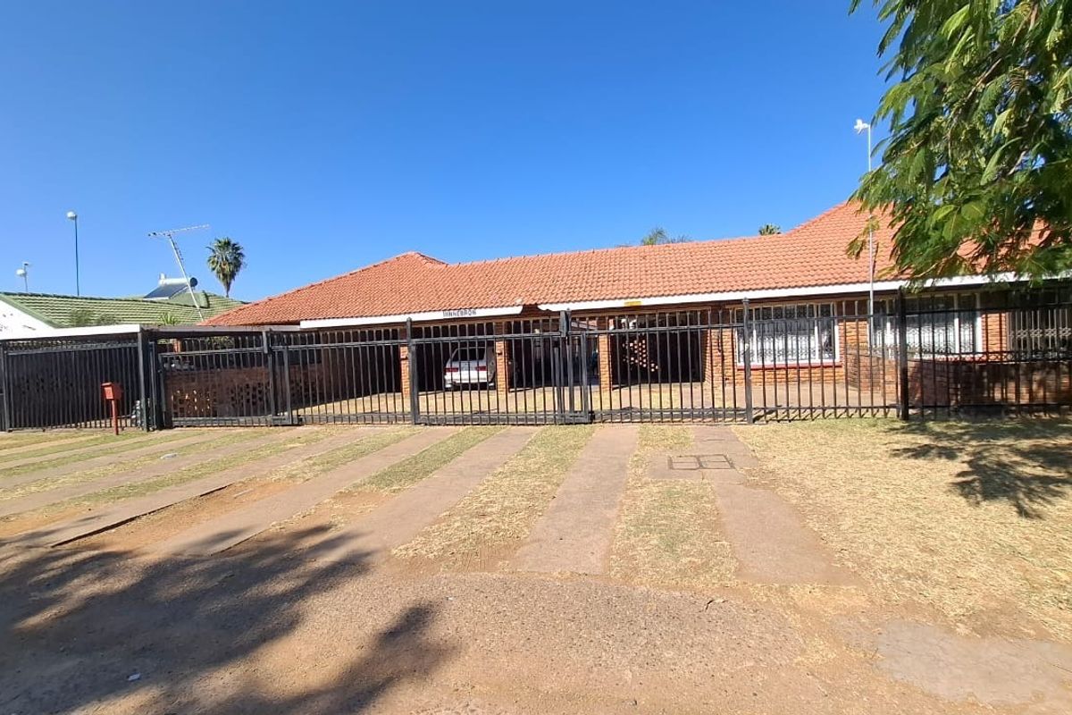 Front elevation of a single-story brick apartment building featuring a tiled roof, secure metal perimeter fencing, and a paved driveway.