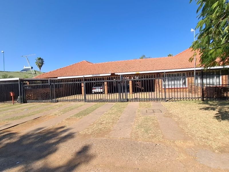 Front elevation of a single-story brick apartment building featuring a tiled roof, secure metal perimeter fencing, and a paved driveway.