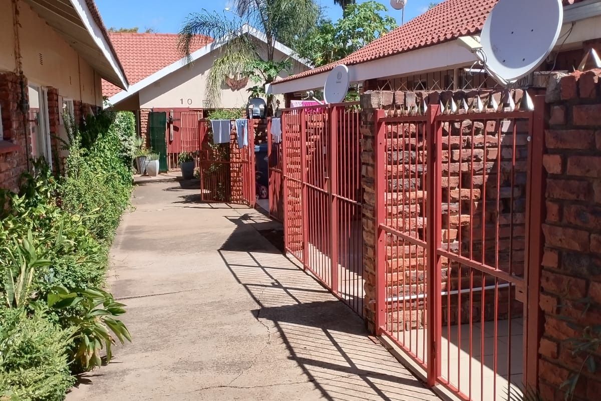 Exterior view of an apartment complex walkway featuring brick boundary walls, metal security gates, and concrete paving between residential units.