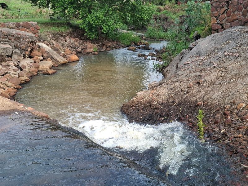 A natural stream flowing over a concrete spillway, flanked by stone retaining walls and rural landscape.
