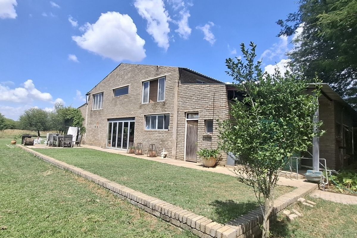 Rear view of a double-story exposed brick small holding residence featuring a paved patio area, sliding glass doors, and expansive surrounding garden space.