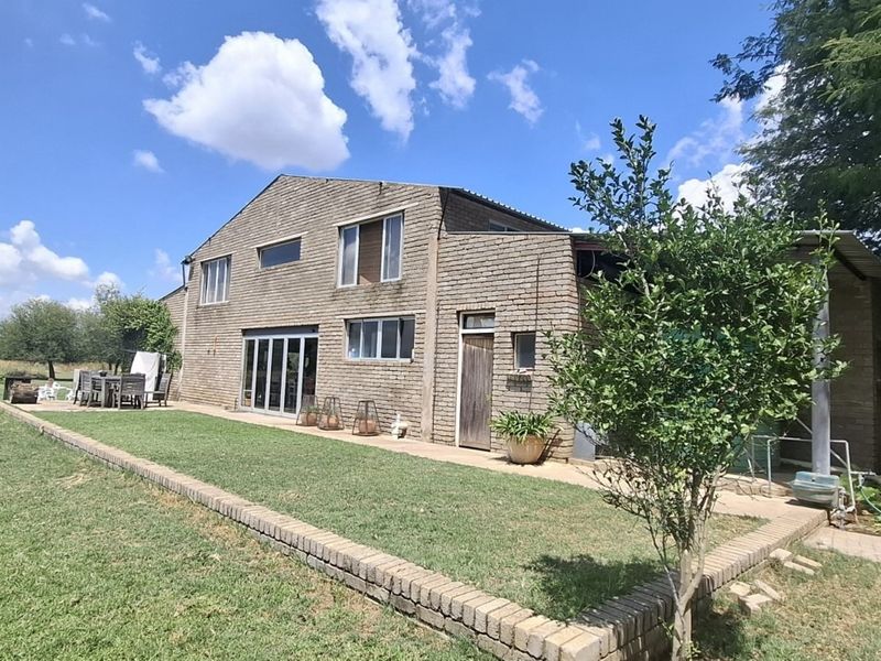 Rear view of a double-story exposed brick small holding residence featuring a paved patio area, sliding glass doors, and expansive surrounding garden space.
