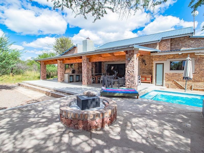 Spacious rear patio featuring a built-in braai and pizza oven, overlooking a swimming pool and a stone-built boma fire pit area.