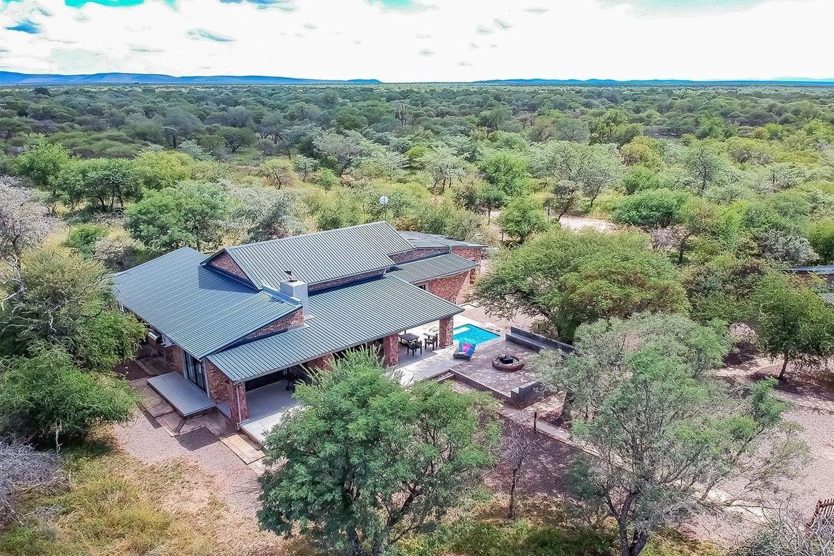 Aerial view of a modern brick house with a metal roof, featuring a swimming pool, covered patio, and outdoor fire pit in a natural setting.