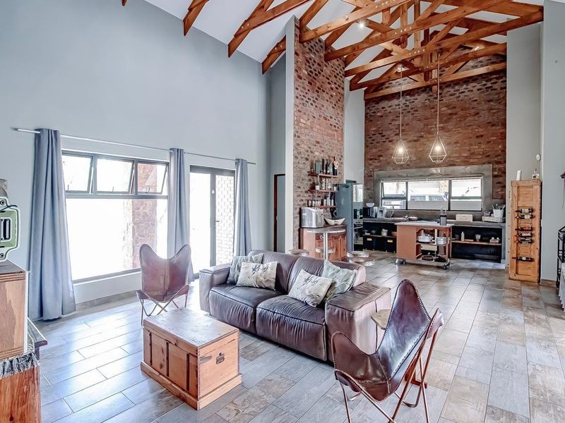 Spacious open-plan living and kitchen area featuring high vaulted ceilings with exposed timber trusses, a striking brick feature wall, and modern tiled flooring.