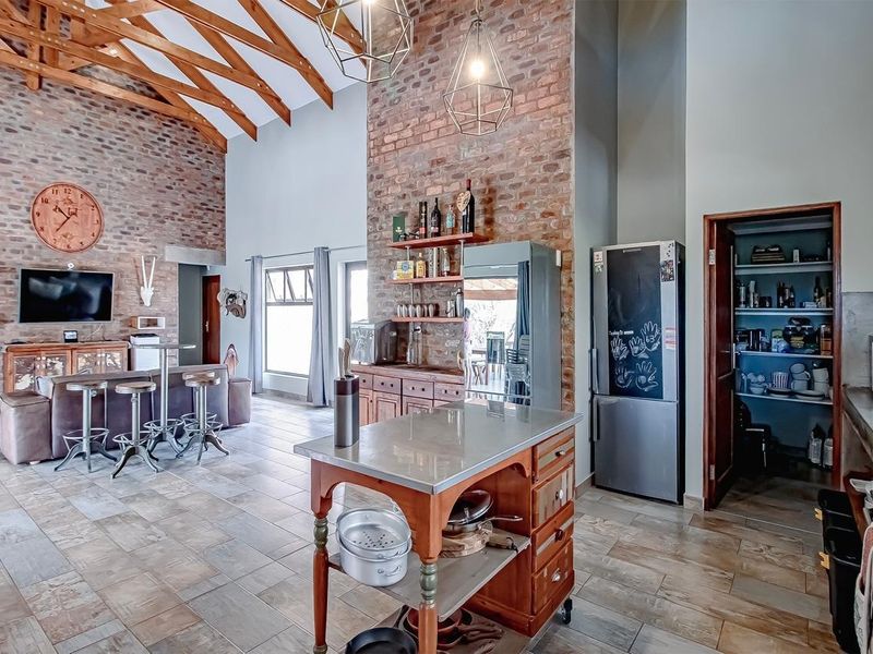 Spacious open-plan living area featuring high vaulted ceilings with exposed timber trusses, brick feature walls, and a functional kitchen with an adjoining walk-in pantry.