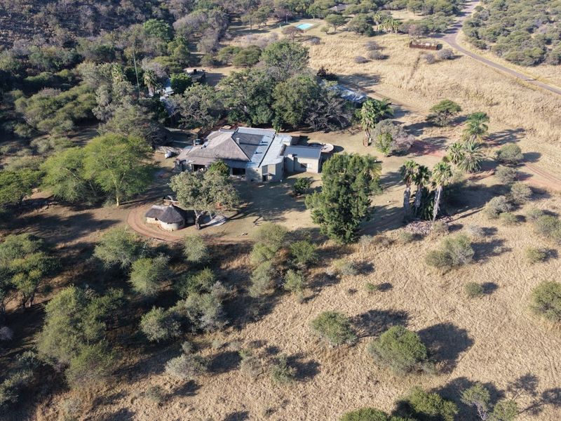 Aerial view of a lodge property featuring a thatched roof main building, swimming pool, and extensive natural grounds within a rural setting.