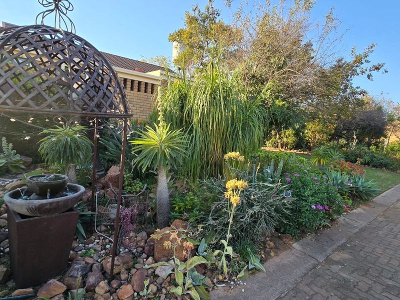 A well-maintained front garden featuring established plants, a decorative metal gazebo structure, a water feature, and a paved driveway area.