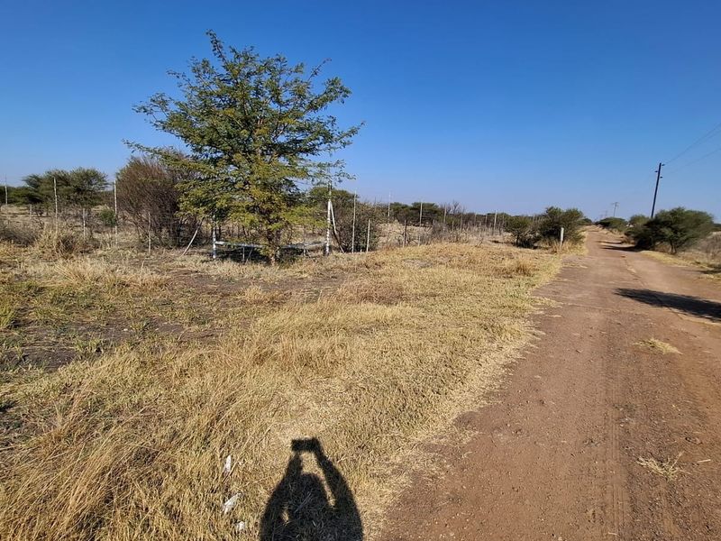 A view of a dirt road bordering a vacant residential plot with wire fencing and natural vegetation.