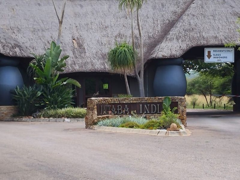 The entrance to Intaba Indlu featuring a distinctive thatched roof gatehouse, stone signage, and a paved driveway leading into the residential estate.