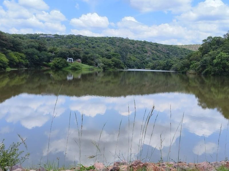 A scenic view of a tranquil lake surrounded by lush natural vegetation and rolling hills under a cloudy blue sky.