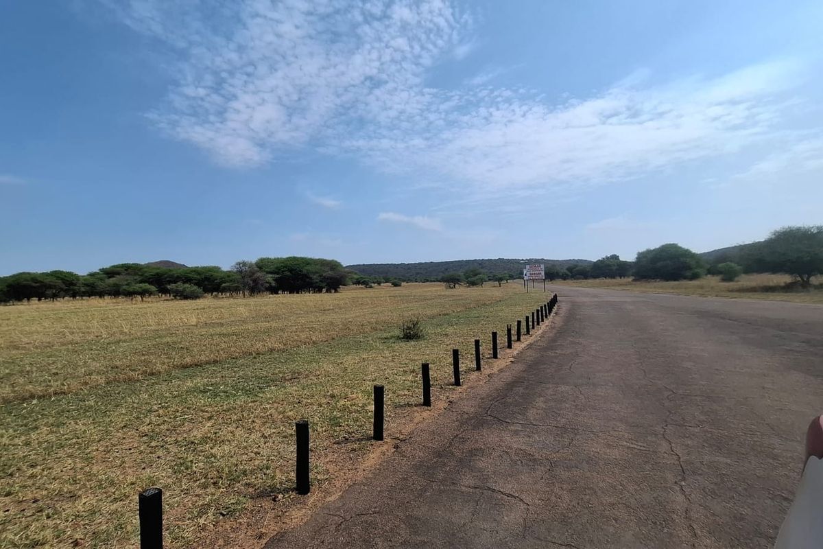A view of a vacant residential land plot featuring an asphalt road frontage and boundary bollards, set against a natural landscape.