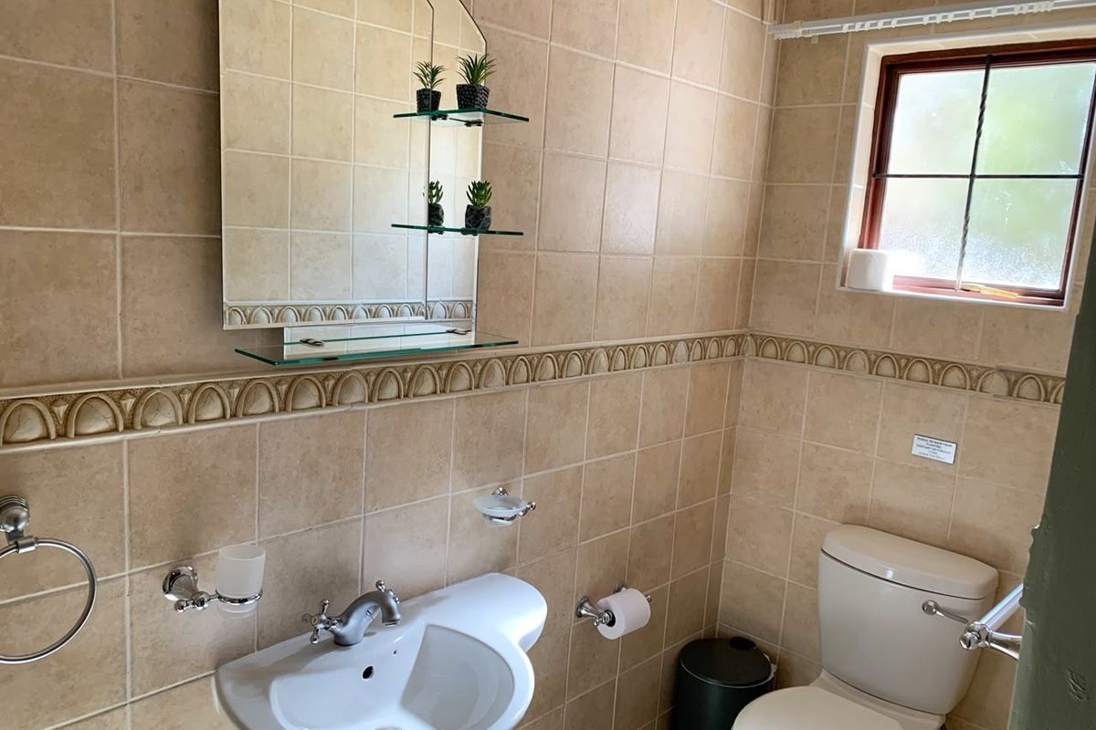 A functional bathroom featuring neutral ceramic wall tiles, a pedestal basin with chrome fixtures, and a wall-mounted mirror with integrated glass shelving.