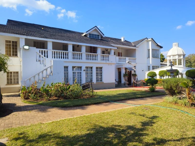 A well-maintained lodge exterior featuring a two-story facade with a balcony, tiled roof, paved walkways, and a landscaped garden with a fountain.