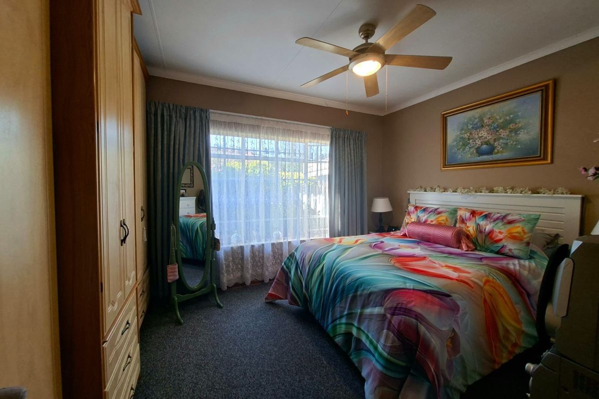 A carpeted bedroom featuring built-in wooden cupboards, a ceiling fan, and large windows with security bars.