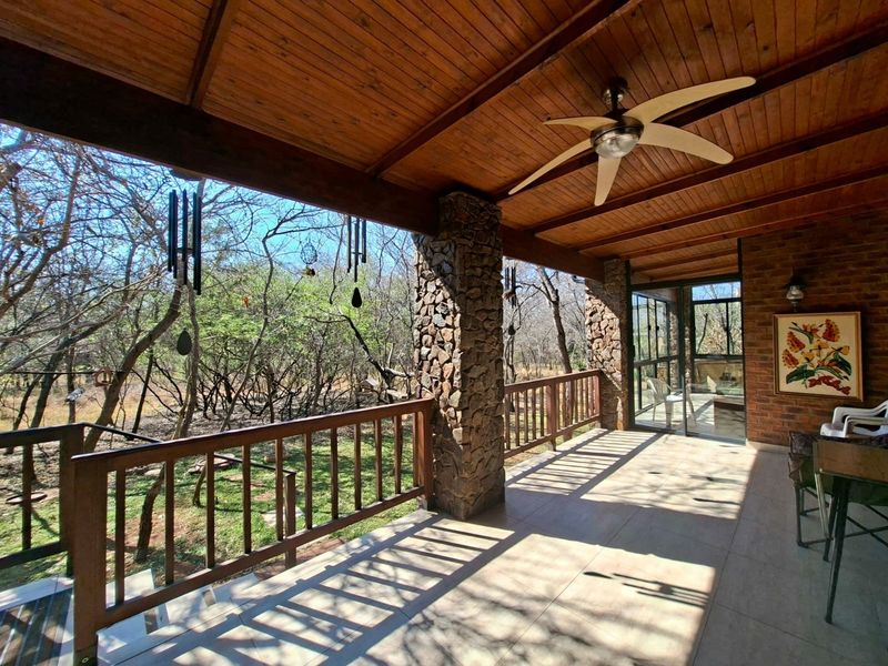 A spacious covered patio featuring a timber ceiling, ceiling fan, stone-clad pillars, and a timber balustrade, overlooking a natural wooded garden area.