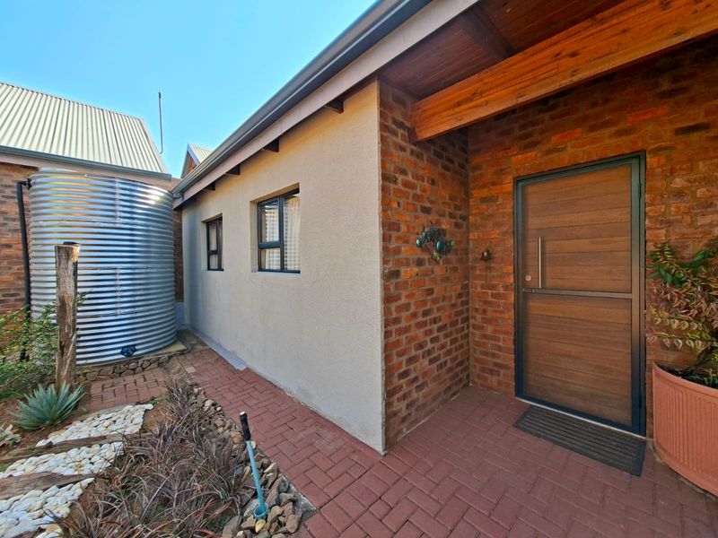 A well-maintained rear exterior area featuring a corrugated water tank, brick paving, and a modern timber entrance door with face brick and plastered wall finishes.