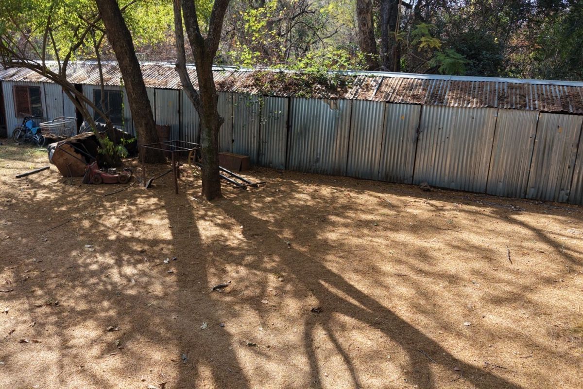 A rear exterior view of the property featuring a long, weathered corrugated metal shed structure situated in a wooded garden area.