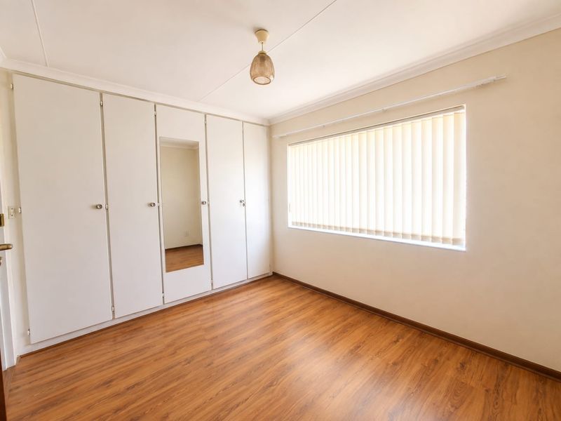 A bright, empty bedroom featuring wood-look laminate flooring, built-in cupboards with a central mirror, and vertical window blinds.
