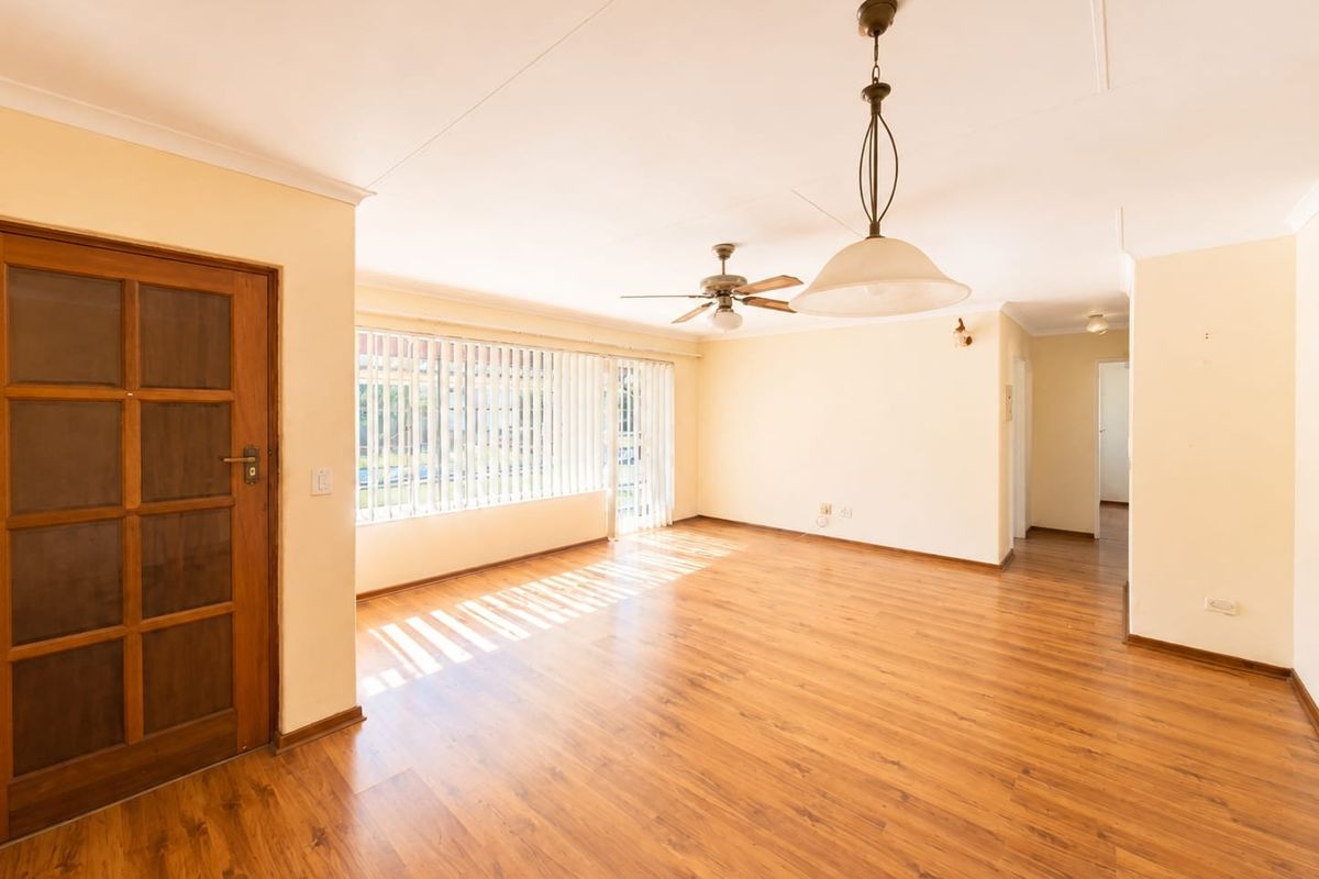 Spacious, light-filled living area featuring warm laminate flooring, a ceiling fan, and large windows with vertical blinds, offering a versatile blank canvas.