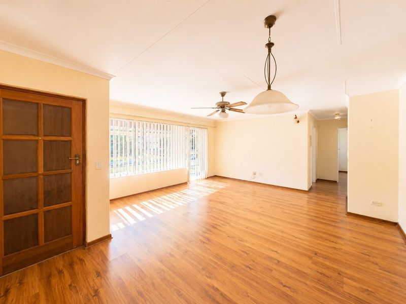 Spacious, light-filled living area featuring warm laminate flooring, a ceiling fan, and large windows with vertical blinds, offering a versatile blank canvas.