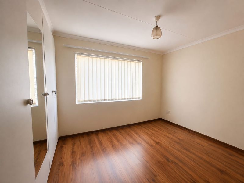 A bright, empty bedroom featuring warm-toned laminate flooring, built-in mirrored wardrobes, and vertical window blinds, offering a clean and neutral space.